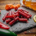 Close-up of honey habanero meat sticks on slate with sliced peppers, lemon slice, and honey bottle.