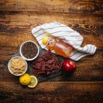 Honey pepper beef jerky on slate with honey, black pepper, brown sugar, lemon, and apple on rustic wood table.