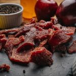 Close-up of Glenwood Original beef jerky pieces on slate with peppercorns, lemon slices, and apples in background.