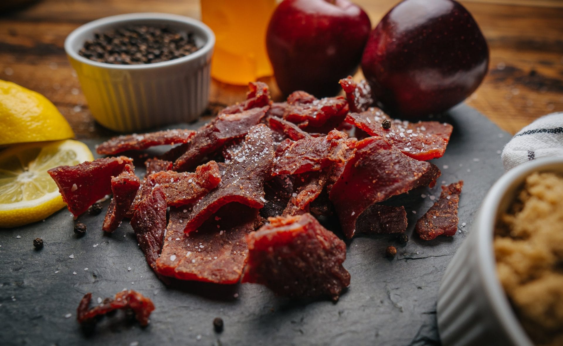 Original-2 Close-up of Glenwood Original beef jerky pieces on slate with peppercorns, lemon slices, and apples in background.