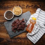 Peppered beef jerky on slate with bowls of black pepper and brown sugar plus honey bear bottle on towel.