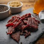 Glenwood Peppered beef jerky on slate with cracked pepper, brown sugar bowls, and honey in background.