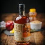 Bottle of bourbon whiskey on a rustic wooden table with honey, apple, and spices blurred in the background.