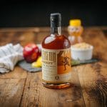 Bottle of bourbon whiskey on a rustic wood table with honey, apple, and lemon blurred in the background.