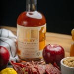 Bottle of bourbon whiskey with beef jerky, lemon, apples, and brown sugar arranged on a rustic table.