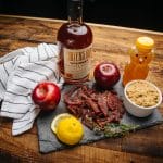 Bottle of bourbon whiskey with honey, apples, lemon, brown sugar, and beef jerky arranged on rustic wood table.