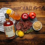 Bottle of bourbon whiskey with honey, apples, lemon, brown sugar, and beef jerky on rustic wood table.