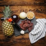 Teriyaki beef jerky on slate with pineapple, soy sauce, brown sugar, onion, garlic, and towel on wood table.